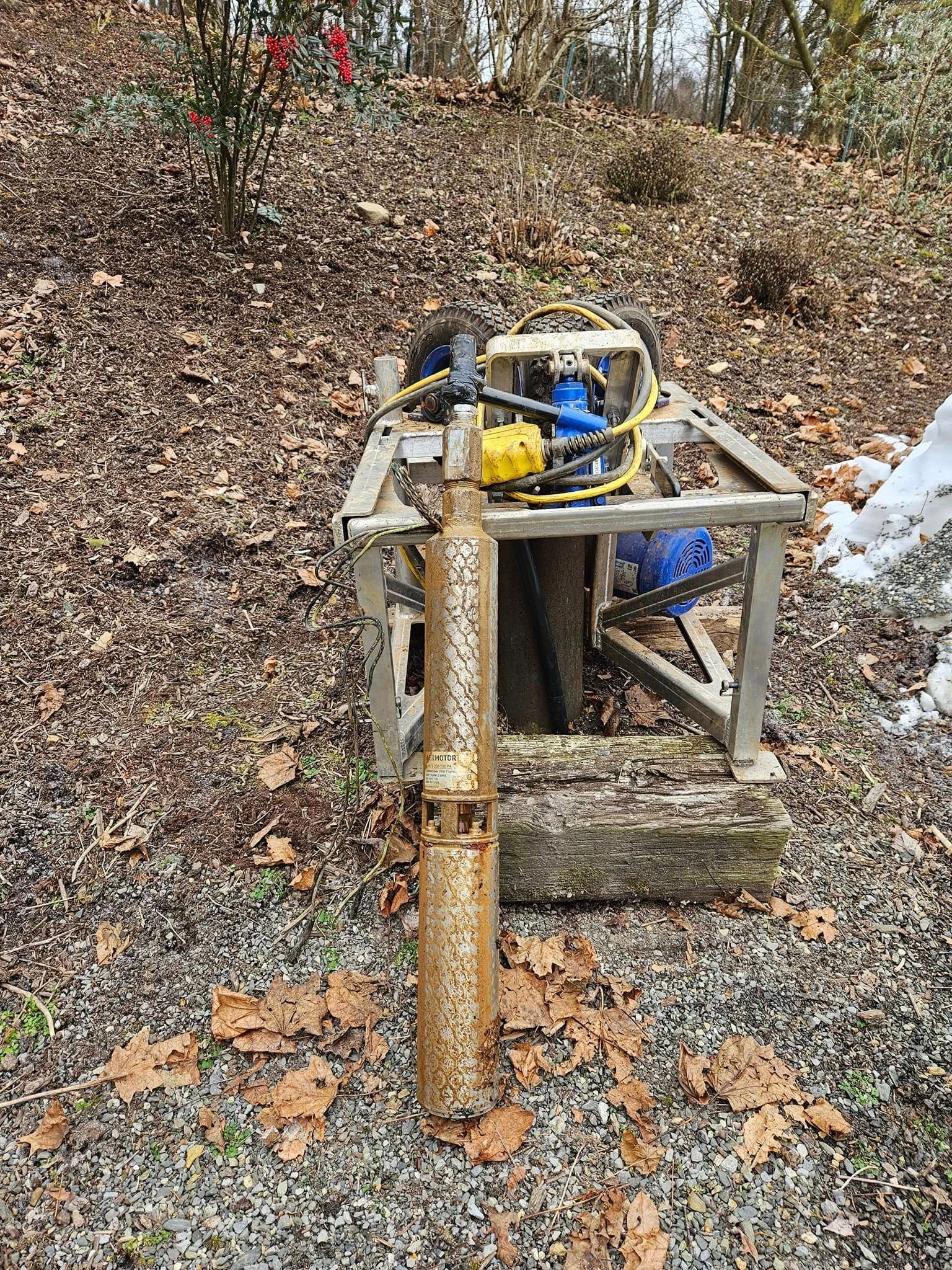 Rusty, cylindrical well equipment with a metal frame and hoses, set in a wooded area with leaves.