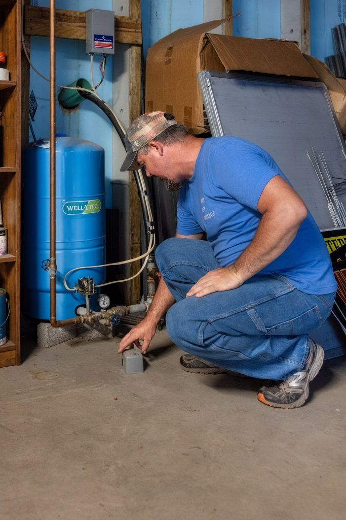 Man examining a water tank in a basement; blue tank, copper pipes, concrete floor, boxes in background.