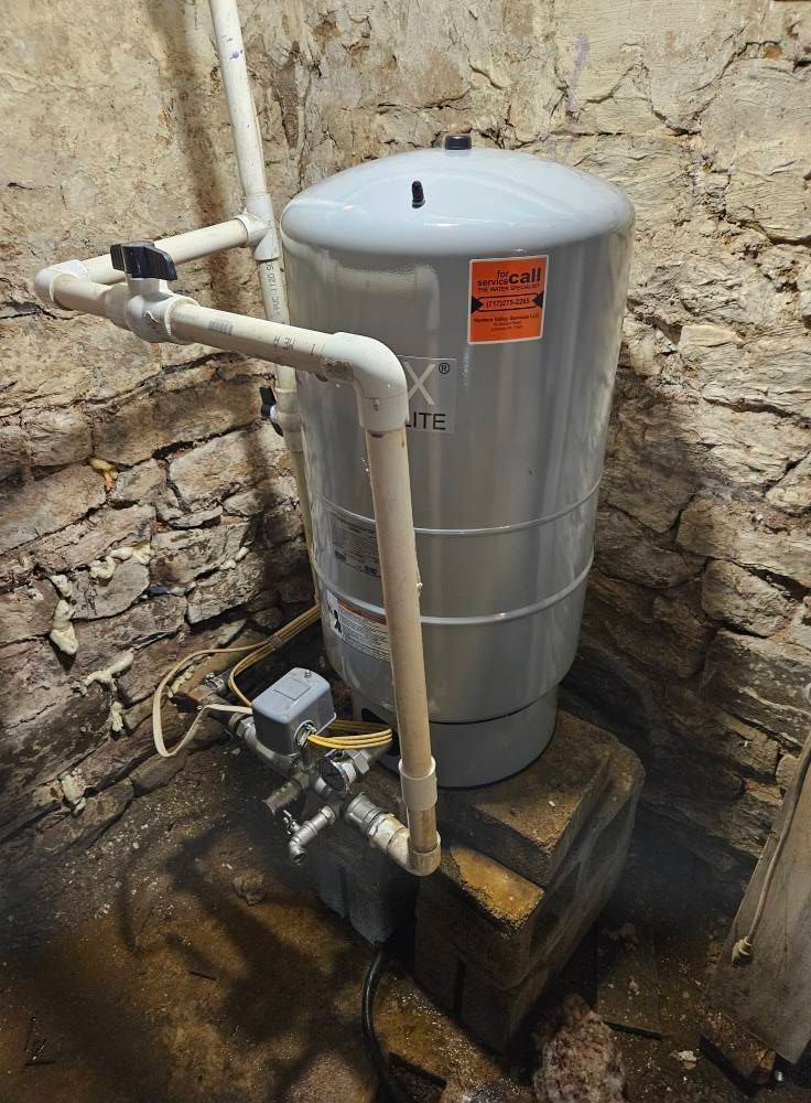 Water pressure tank in a stone-walled basement. White pipes connect to the gray tank.
