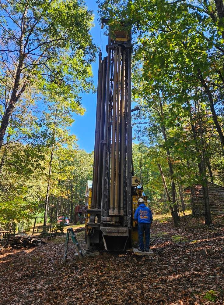 Drilling rig in a wooded area; worker in blue jacket, trees, clear blue sky.
