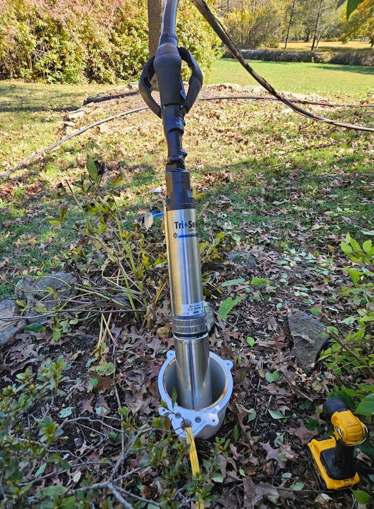 A well pump, installed in the ground, with a yellow power cord, and a cordless drill.