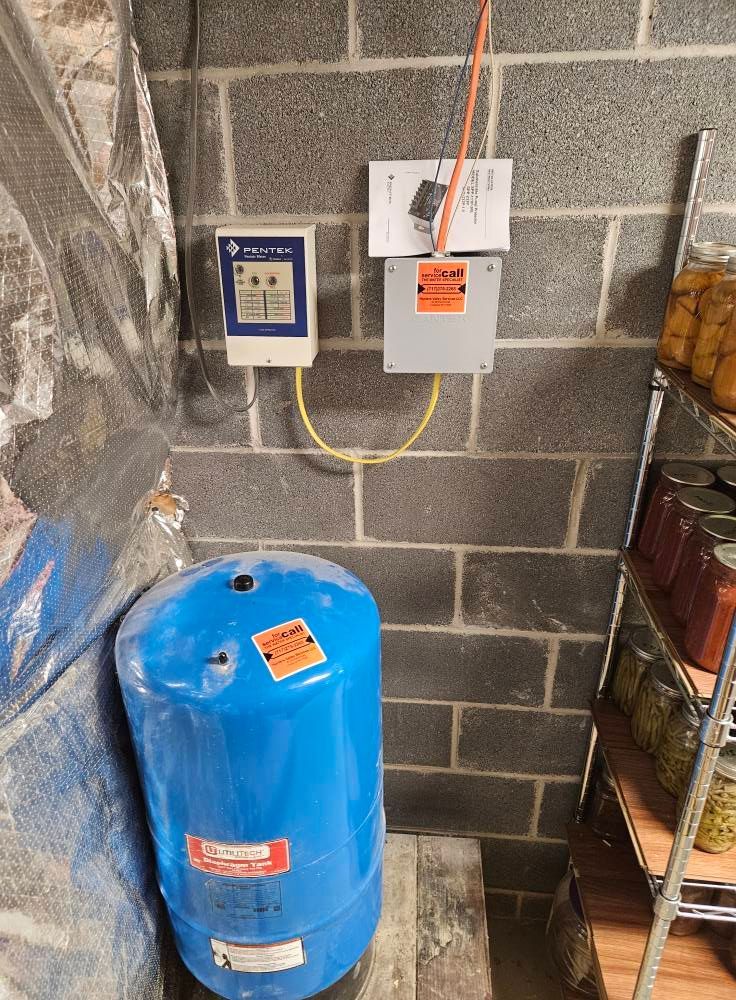 Blue water tank and electrical components mounted on a cinder block wall, with shelving of canned goods to the right.