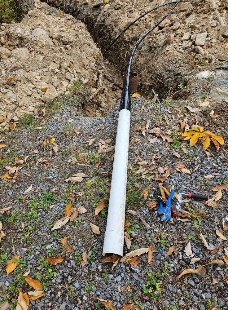 Black cables emerge from a white protective sleeve in a trench. Blue handled tool and knife nearby.