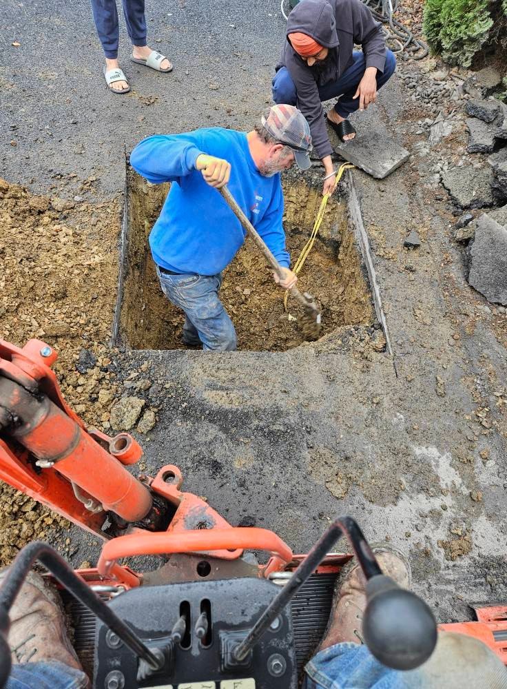People digging in a square hole in the ground, an excavator in the foreground.