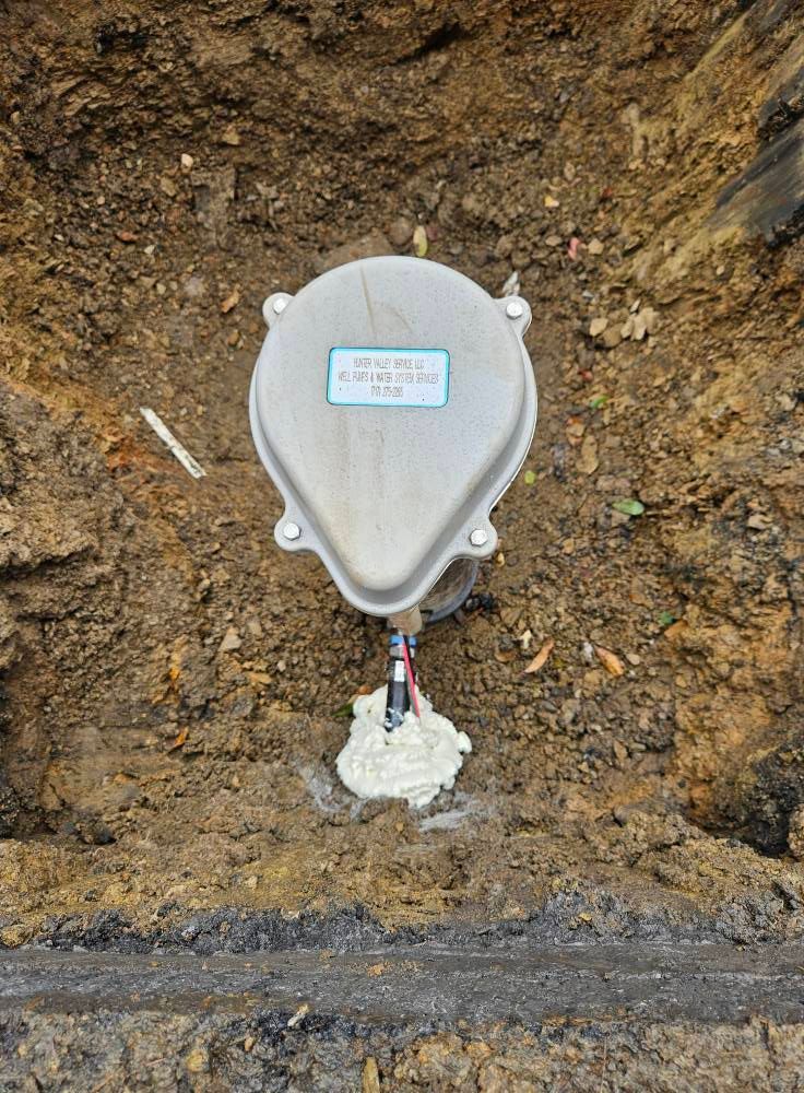 Gray electrical junction box in a dug-out area, sealed with white foam. The box has a label and wires.