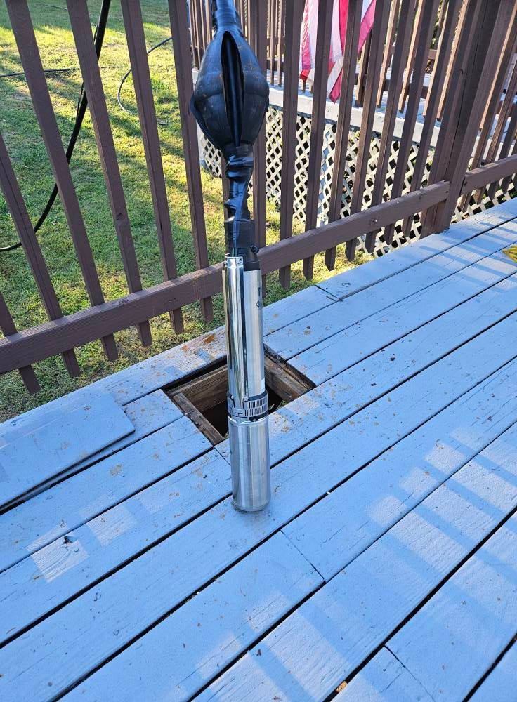 A well pump is being lowered into a square hole in a wooden deck.