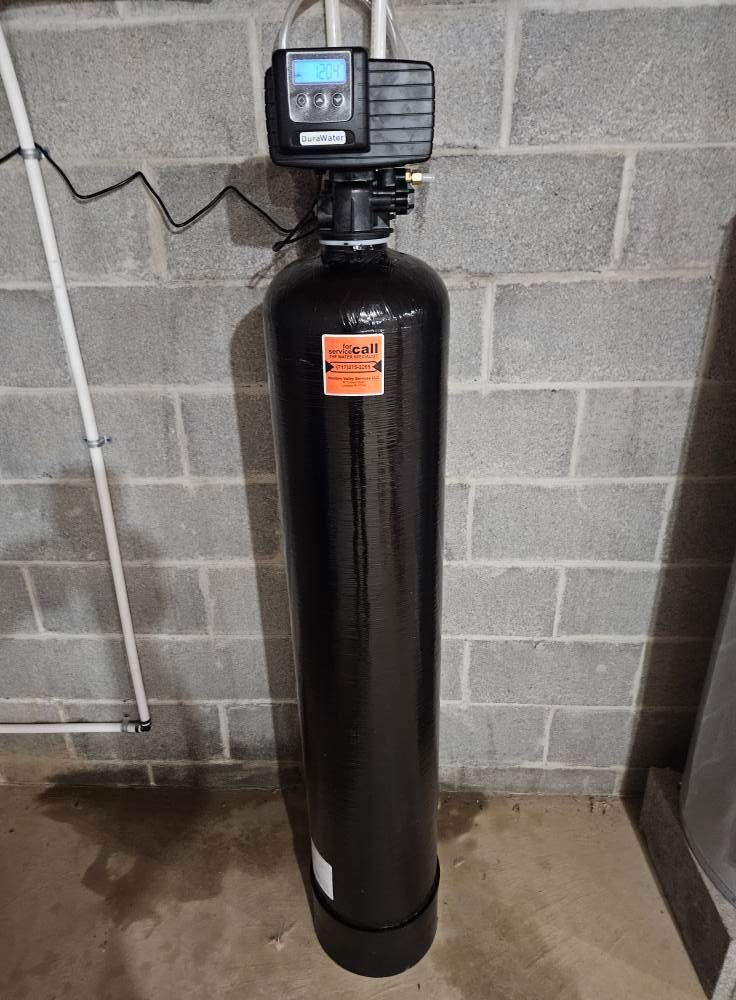 A black water softener tank with a digital head is against a gray brick wall.