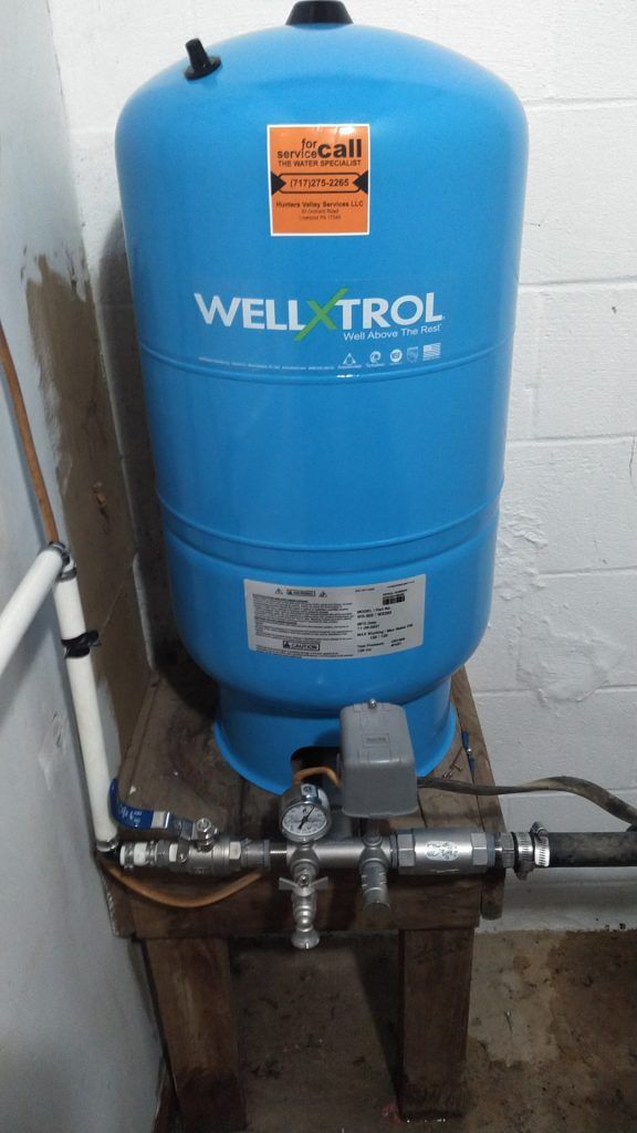 Blue Well-X-Trol water pressure tank on wooden stand with connected pipes and gauges in a basement.