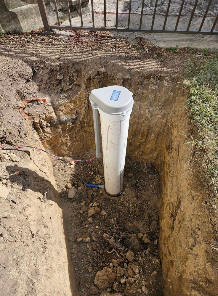 A white cylindrical water filter in a rectangular trench in a yard, with dirt and grass visible.
