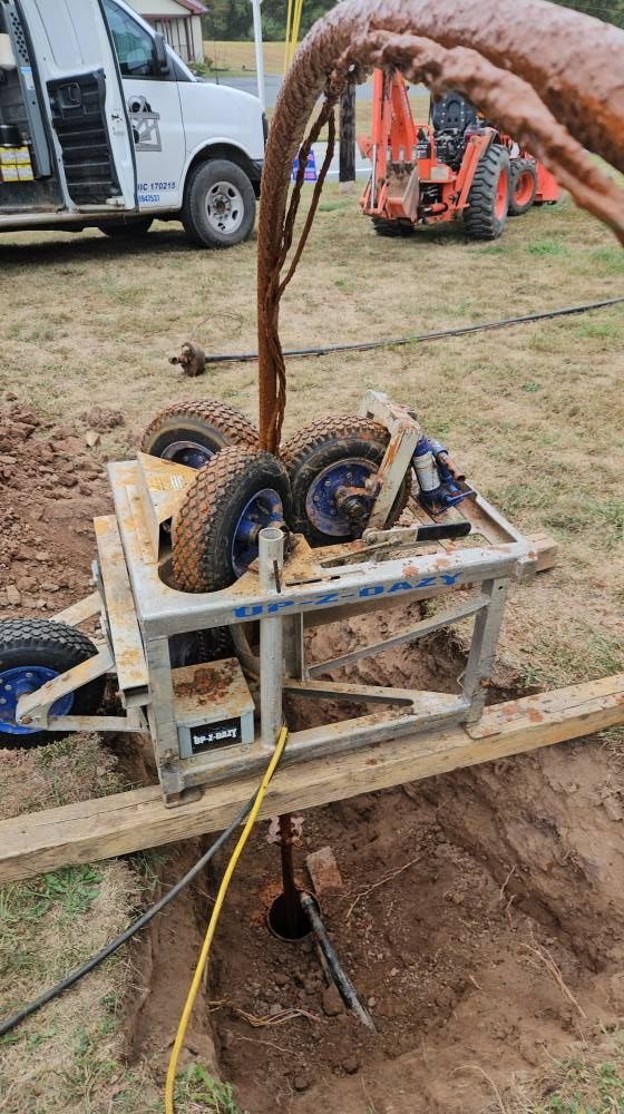 Well drilling rig near a hole in the ground, with a white van and orange tractor in the background.