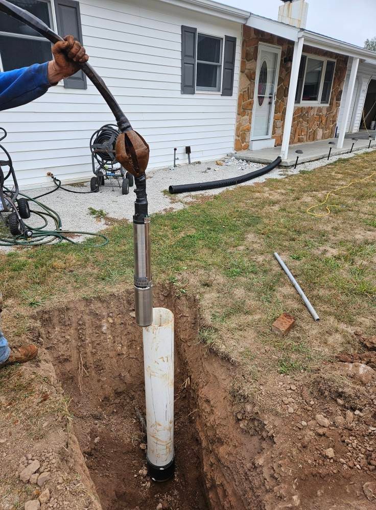 A person lowering a pump into a well casing in a trench outside a house.