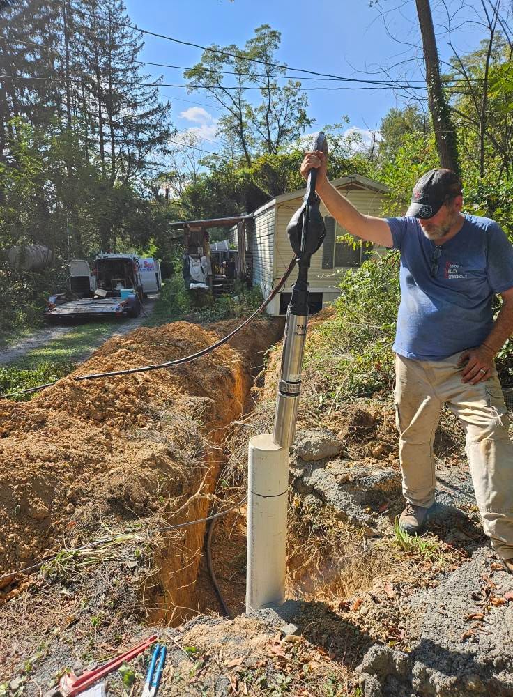 Man operating machinery to install a post in a trench. Outdoor setting, brown dirt, trees, and small house.