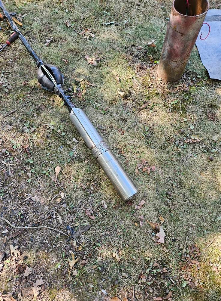 Silver well-drilling tool lying on grass next to a metal casing in an outdoor setting.