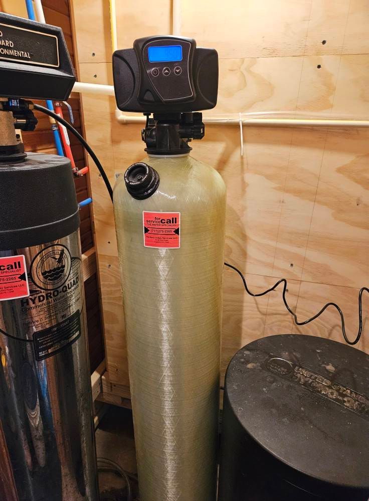 Water softener system with control panel, cylindrical tank, and black brine tank in a utility room.