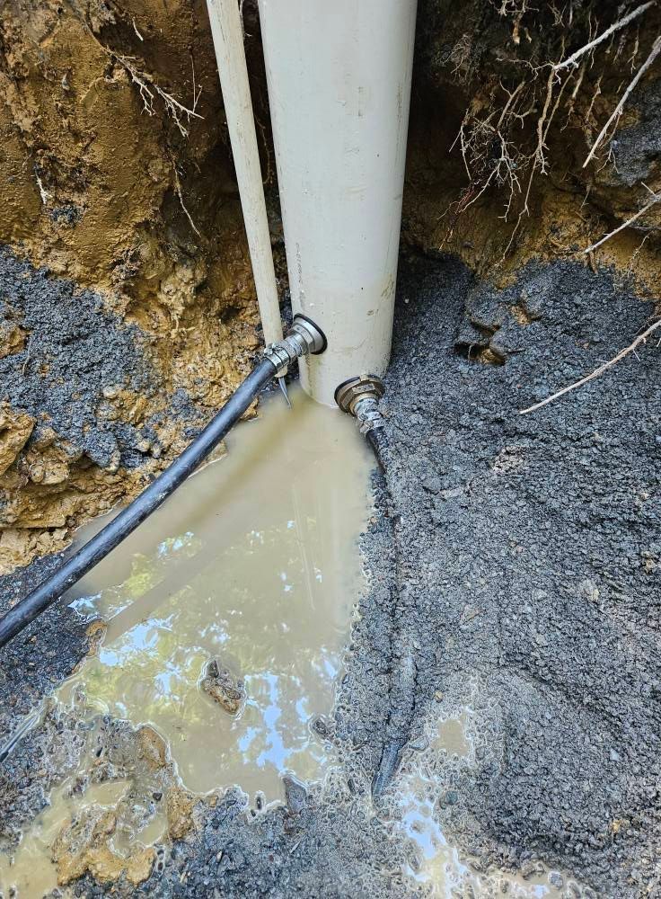 White pipe with attached hoses leaking water into a muddy trench.