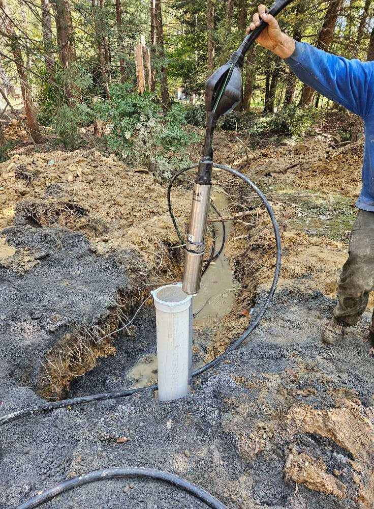 Man operating a well pump in a muddy trench; equipment, water, and tubing visible.