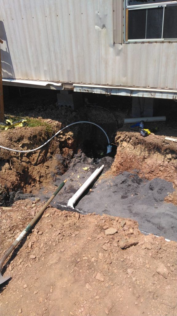 Construction site with exposed pipes and wiring near the foundation of a mobile home. Soil and tools are visible.