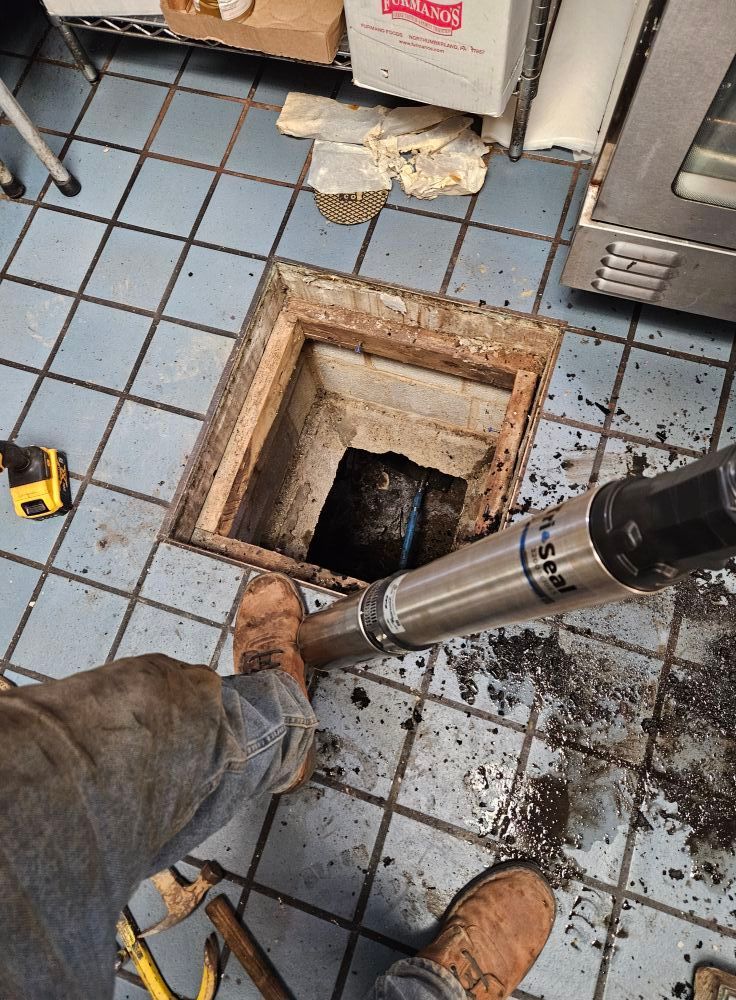 Person using a tool to work on a floor drain in a tiled commercial kitchen.
