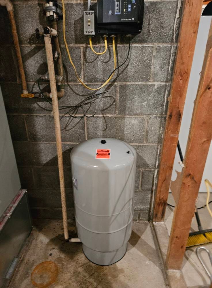 Water pressure tank in a basement, connected to pipes and electrical components against a cinder block wall.