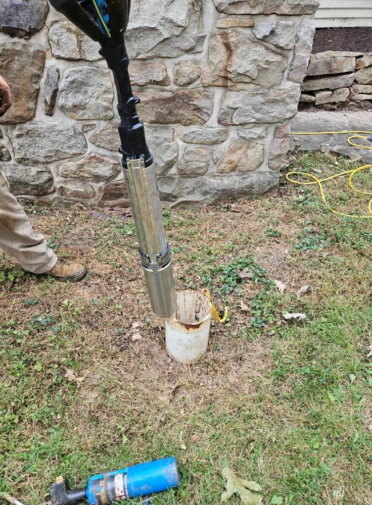 Person lowering a well pump into a well casing in a grassy yard, near a stone wall.