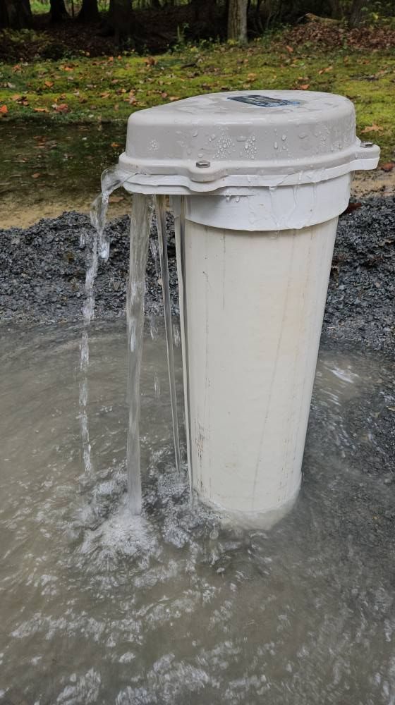 A white cylindrical trash can overflowing with water, creating a puddle on gray gravel.