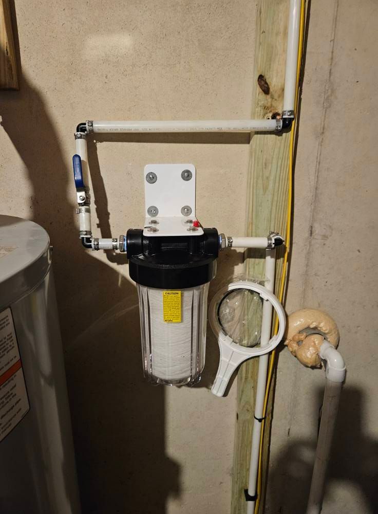 Water filtration system mounted on a wall in a basement, with a clear filter housing, white piping, and a wrench.