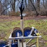 A portable drilling rig with blue wheels, in a grassy outdoor setting.