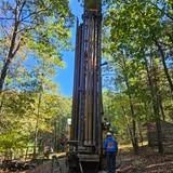 Drilling rig in a wooded area with a worker in blue standing nearby.