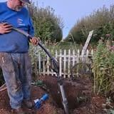 Man digging a trench with a shovel near a white picket fence in a garden.