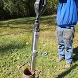 Person in blue jacket and jeans holding a water well pump, outdoors on grass.