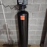 A tall black water softener tank with a digital display attached to a gray cinder block wall.
