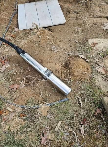 A silver submersible well pump laying on dirt and leaves with a black cable attached.