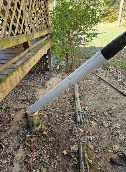 PVC pipe angled against a wooden post, near a deck and foliage.