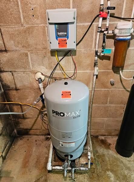 Water pressure tank with control box and filter against a cinder block wall in a basement.