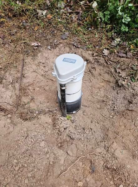 A cylindrical white and grey device on dirt ground, possibly a monitoring station in a natural setting.