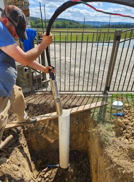 Man lowers pipe into a rectangular trench, using tool attached to black hose; near a metal fence.
