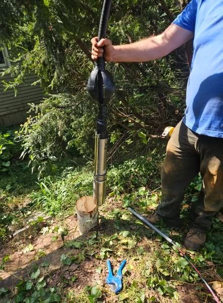 Person holding a water well pump, outdoors. The pump is by a log; blue gloves are nearby.