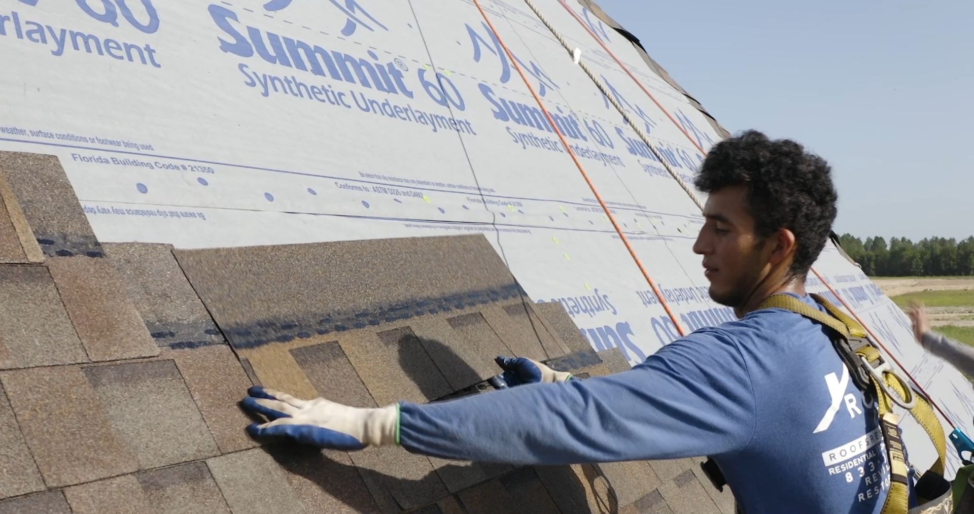 Roofer installing shingles on a roof, wearing safety gear. He is focused, working in a sunny environment.