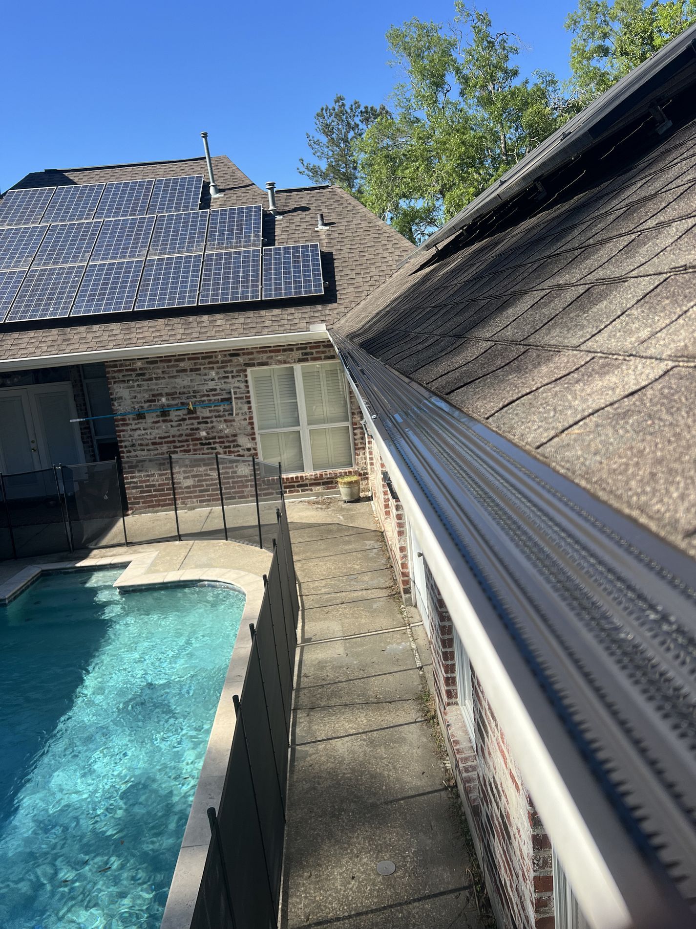 A home's roof with solar panels and a gutter. A brick exterior with a pool below. Sunny day.