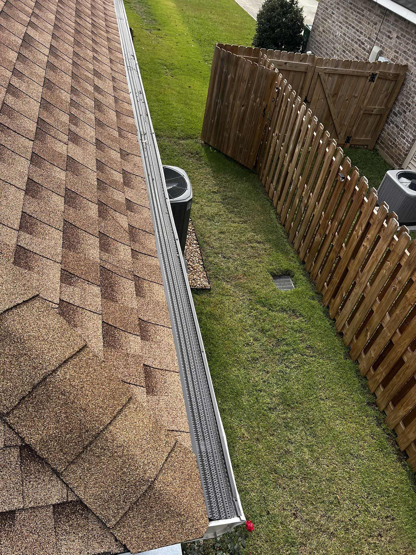 Brown shingled roof with a metal gutter, bordering a green lawn and wooden fence.