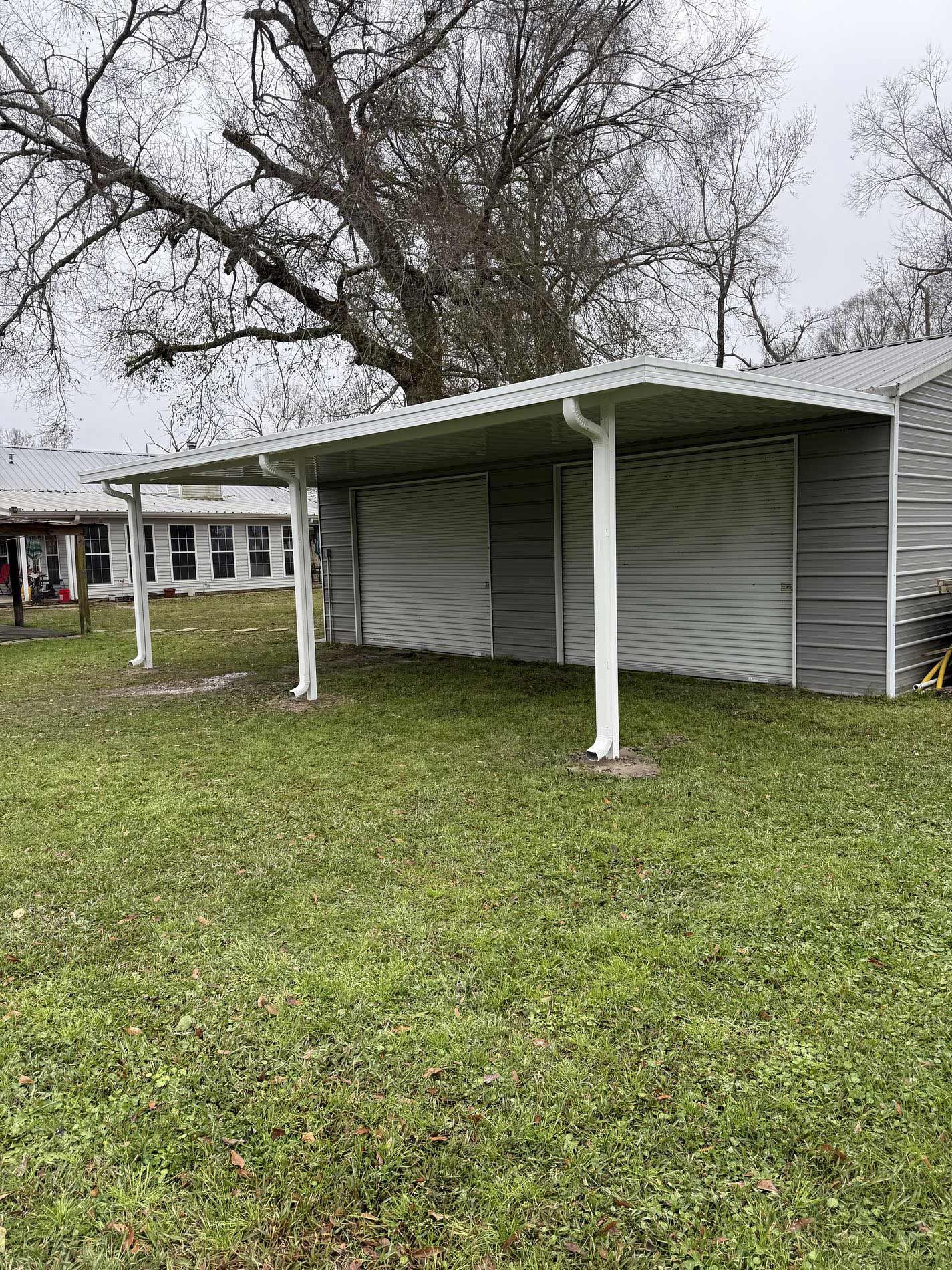 A white-roofed carport attached to a gray-sided building. It has white support beams on a grassy lot.