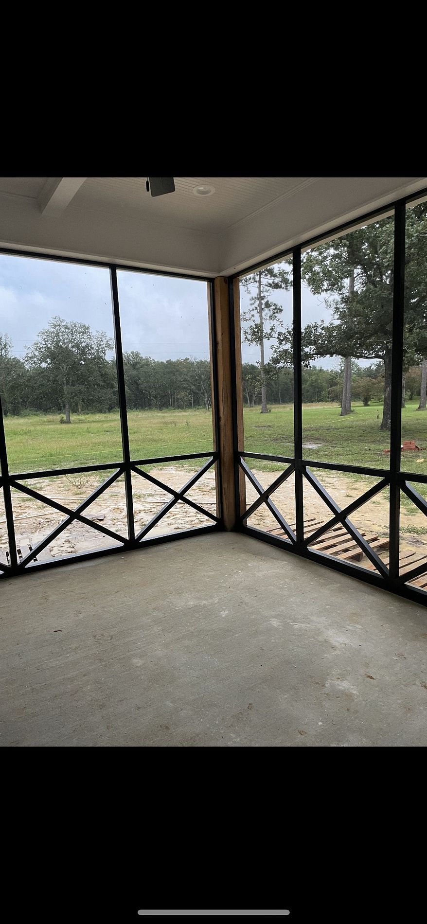 A screened porch with a view of a grassy field and trees. The porch has a concrete floor and decorative railing.