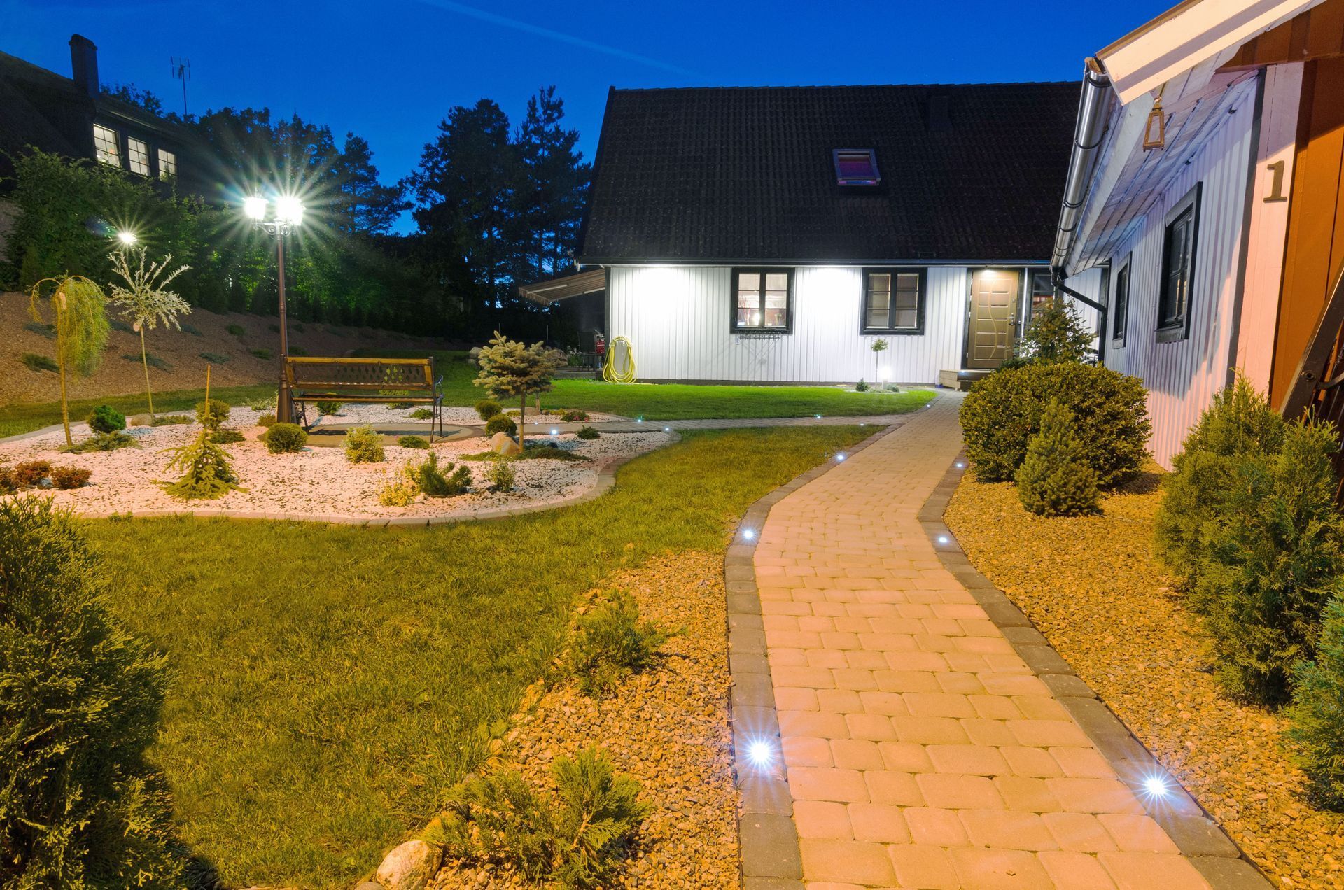 Nighttime view of a lit garden path leading to a small house with a paved yard and grass.