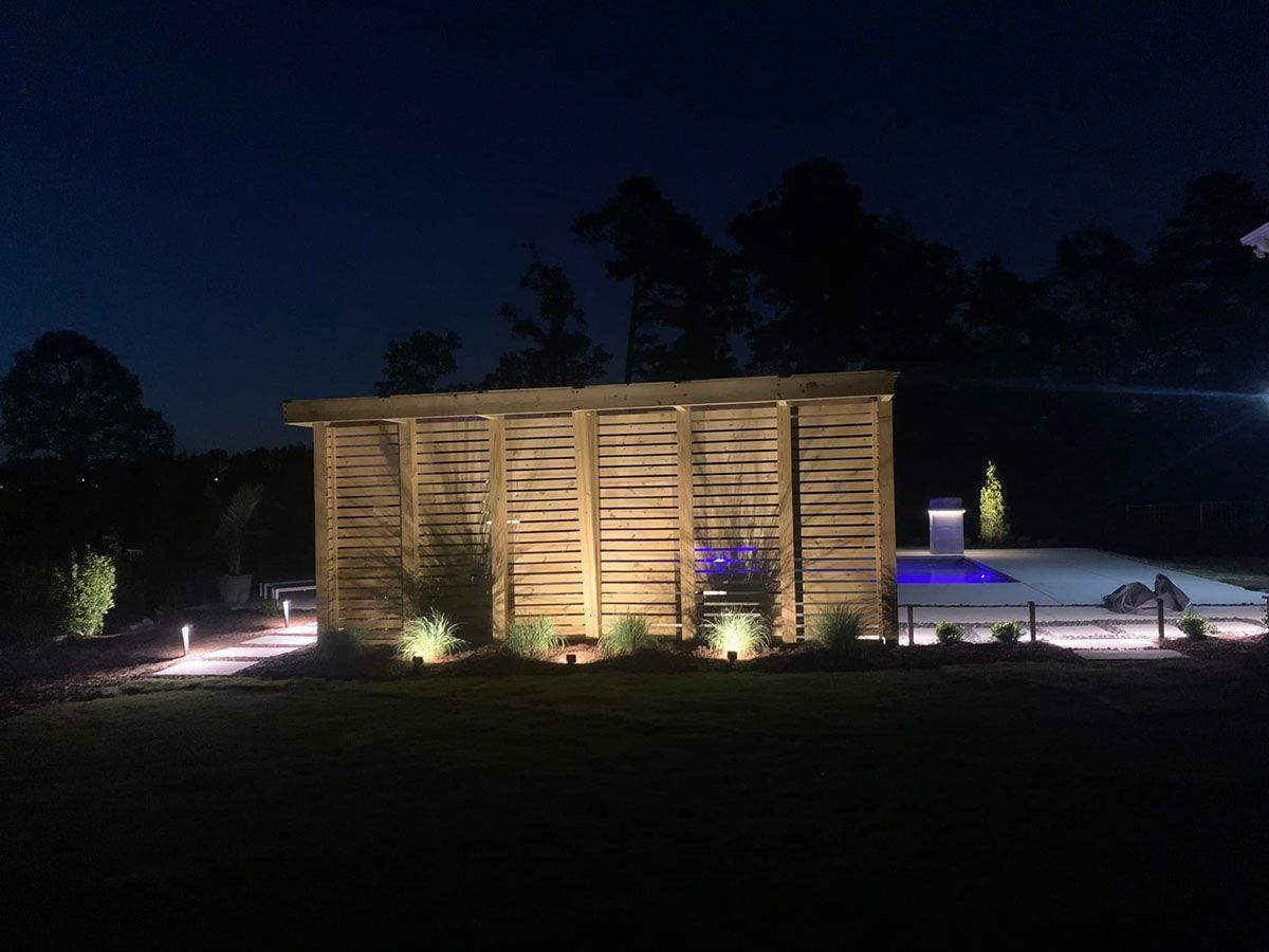 Lit wooden pavilion beside a glowing pool at night, with pathway lights and dark sky behind