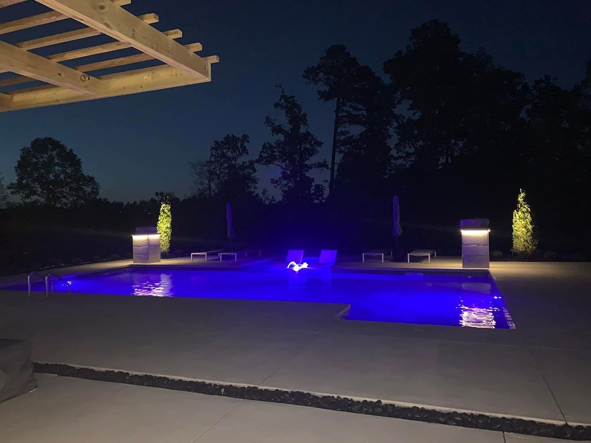 Nighttime backyard pool with blue-purple lighting, patio, and silhouetted trees in the background