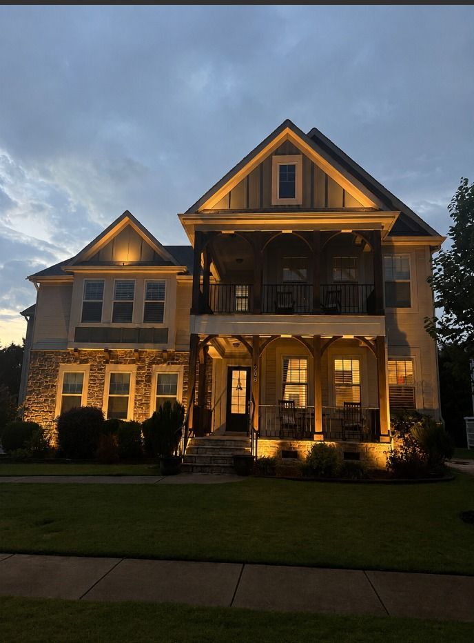 Large two-story house with warm exterior lights at dusk under a cloudy sky