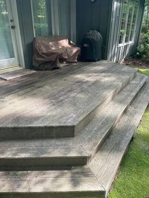 Weathered wooden deck with two steps, a covered chair, and sliding glass doors beside a grassy yard.