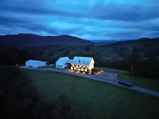 White house glowing at dusk on a rural hillside, with mountains and cloudy blue sky in the background