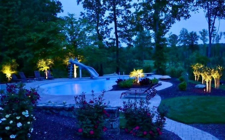 Illuminated backyard pool and patio at dusk, with landscaped flowers, trees, and lounge chairs.
