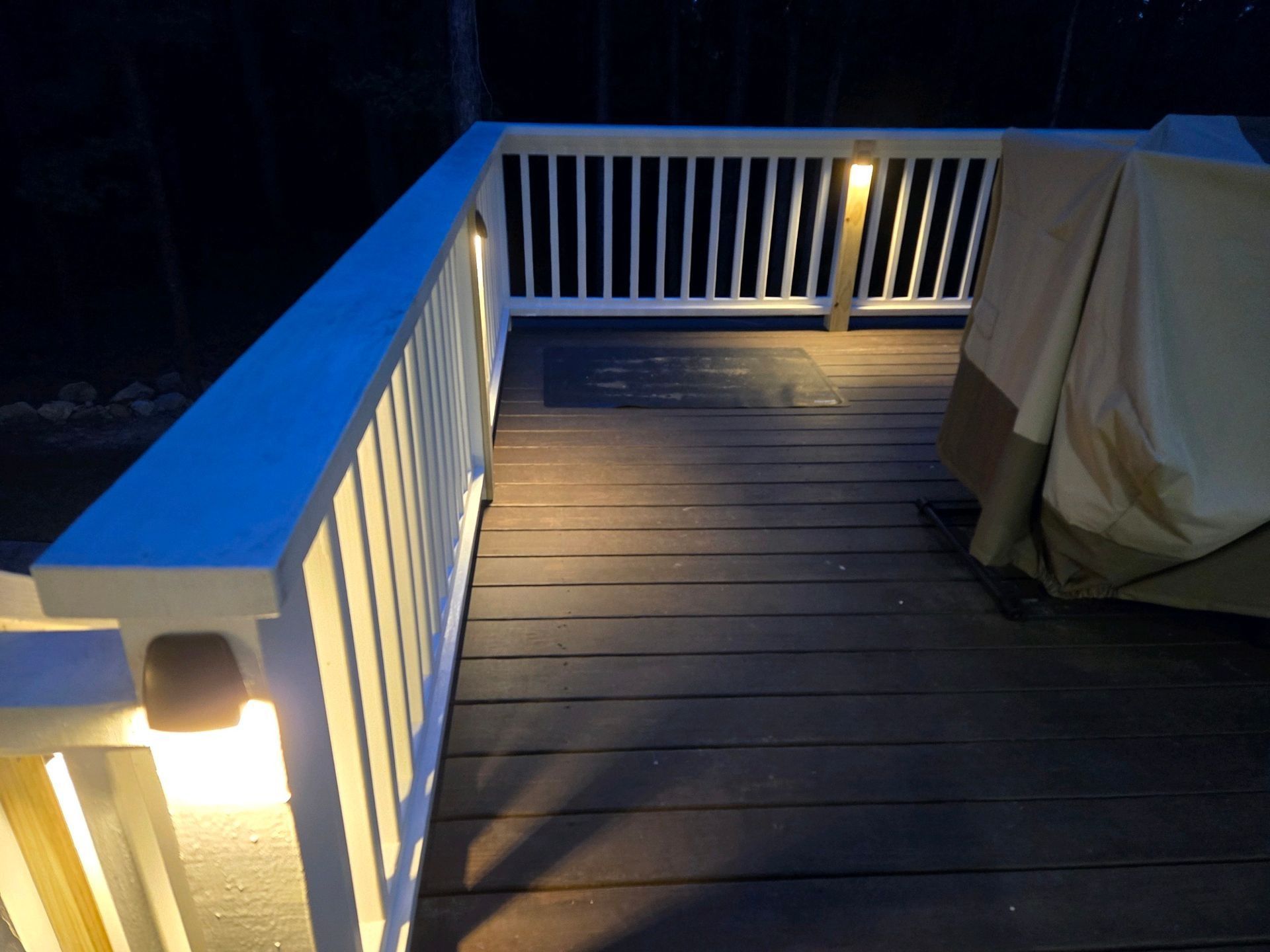 Lit wooden deck with white railing at night, covered chair on the right, and a warm porch light glowing.